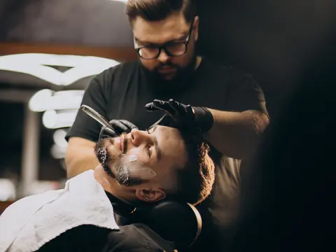 Barbero de anteojos afeitando con navaja a un cliente recostado en el sillón