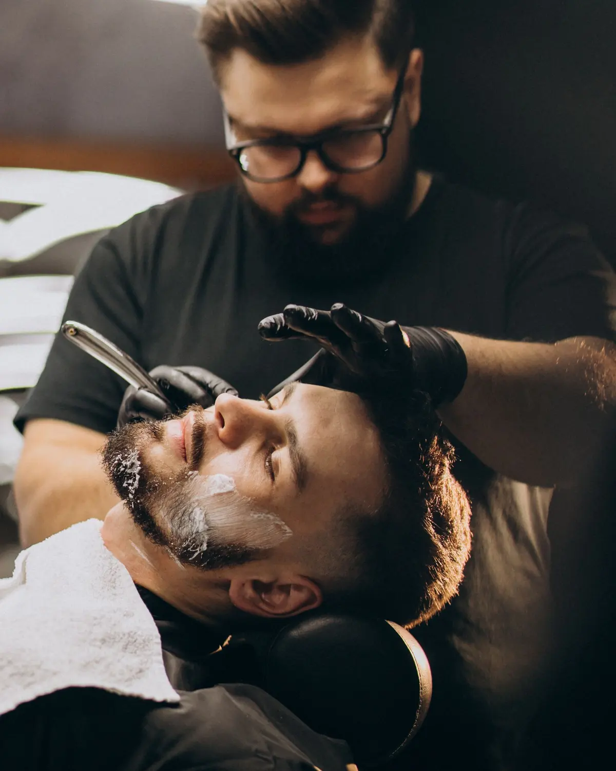 Barbero de anteojos afeitando con navaja a un cliente recostado en el sillón de la barbería