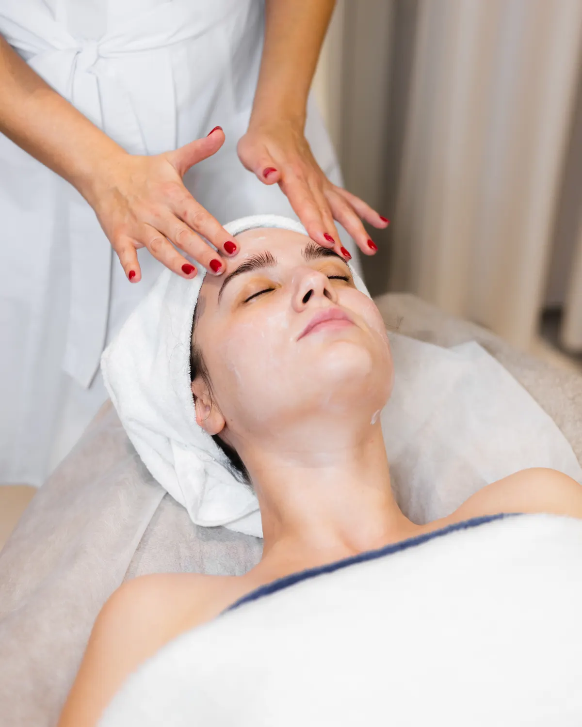 Esteticista en bata blanca masajeando la frente de una clienta con toalla en la cabeza, recostada en una camilla de spa