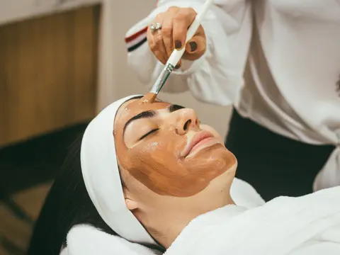 Esteticista aplicando una mascarilla con pincel a una clienta con toalla en la cabeza