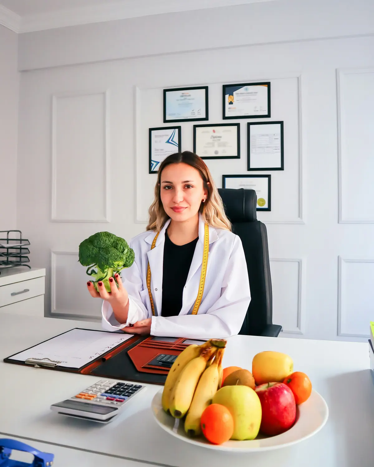 Nutricionista con guardapolvo blanco sosteniendo un brócoli en su consultorio, con una frutera sobre el escritorio